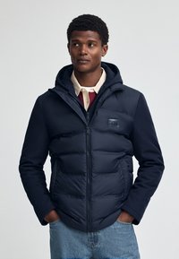 Marinblå vadderad jacka med dragkedja och huva, som har en logotypapplikation. Slät textur med kontrasterande tyg på ärmarna. Enkel design, funktionell stil.