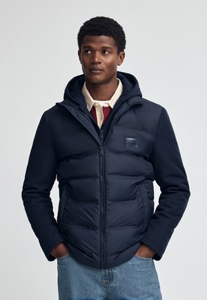 Marine gewatteerde jas met een rits en capuchon, voorzien van een logo patch. Gladde textuur met contrasterende stof op de mouwen. Eenvoudig ontwerp, functionele stijl.