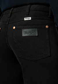 Black denim jeans with a leather patch featuring the "Wrangler" logo, copper rivets, and a textured zigzag pattern on the back pockets.