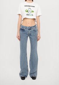 Avskuren vit t-shirt med grafisk design och text, kombinerad med ljusblå, utsvängda jeans med framfickor och knappstängning.