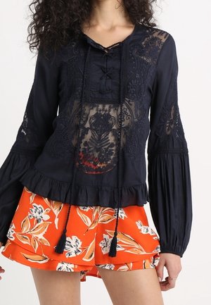 Kobieta ubrana w czarną prześwitującą koronkową bluzkę z długimi rękawami oraz pomarańczowe kwieciste szorty z warstwowymi falbankami.