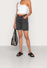 Shorts di jeans neri a vita alta, abbinati a un crop top bianco con spalla scoperta e una borsa a rete nera. Sandali neri a scivolo completano il look.