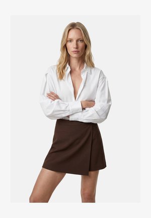 Vaaleahiuksinen nainen, jolla on kädet ristissä, yllään löysä valkoinen nappipaita ja tummanruskea kietaisuhame, seisoo valkoisen taustan edessä.