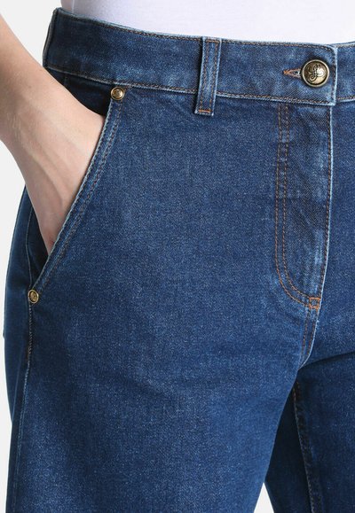 Gros plan d'un jean en denim bleu avec une main dans la poche avant, montrant le bouton métallique et les détails de couture.