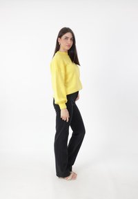 Maglione giallo lavorato a maglia con maniche lunghe e polsini a coste, abbinato a pantaloni neri a vita alta e sandali con tacco di colore chiaro.