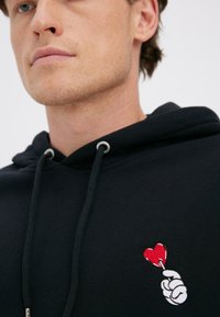 Čierna mikina s kapucňou a sťahovacou šnúrkou, na hrudi vyšívaný motív bielej ruky držiacej červené srdce. Hladká textúra.