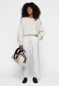 Hellgrauer, taillierter Pullover mit langen Ärmeln, kombiniert mit hellgrauen Jogginghosen. Hält eine beigefarbene Tasche mit schwarzen Streifen und trägt dunkle Sneaker.