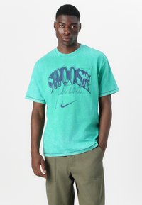Minta zelena bombažna majica z velikim vijoličnim grafičnim znakom "Swoosh" in besedilom "just do it" spredaj. Okroglo ovratnik in kratki rokavi.