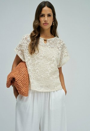 Femme portant un top en dentelle blanche à motif floral et un pantalon blanc ample, tenant une grande pochette orange tissée pliée sous un bras.