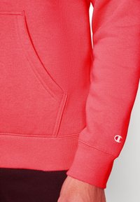 Sudadera de color coral con un bolsillo frontal, puños acanalados y dobladillo. Presenta un pequeño logo blanco en la muñeca. La tela parece suave y texturizada.