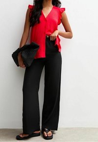 Rode gerimpelde blouse met knopen, gecombineerd met zwarte wijde broek. Het model houdt een gestructureerde zwarte clutch vast en draagt zwarte sandalen.