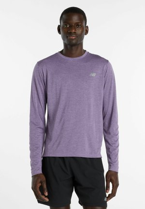 Mand iført en lyse lilla langærmet sportsbluse og sorte shorts, stående mod en hvid baggrund.