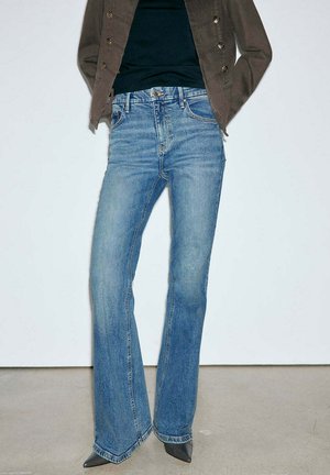 Persona con vaqueros acampanados azul medio, zapatos negros puntiagudos, camiseta negra y chaqueta marrón, de pie con las manos en los bolsillos.