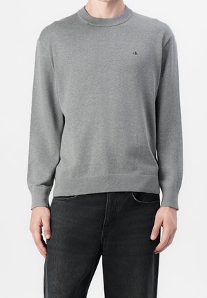 Homme portant un pull gris uni à col rond avec un petit logo "cK" sur la poitrine, associé à un jean noir, debout sur fond blanc.