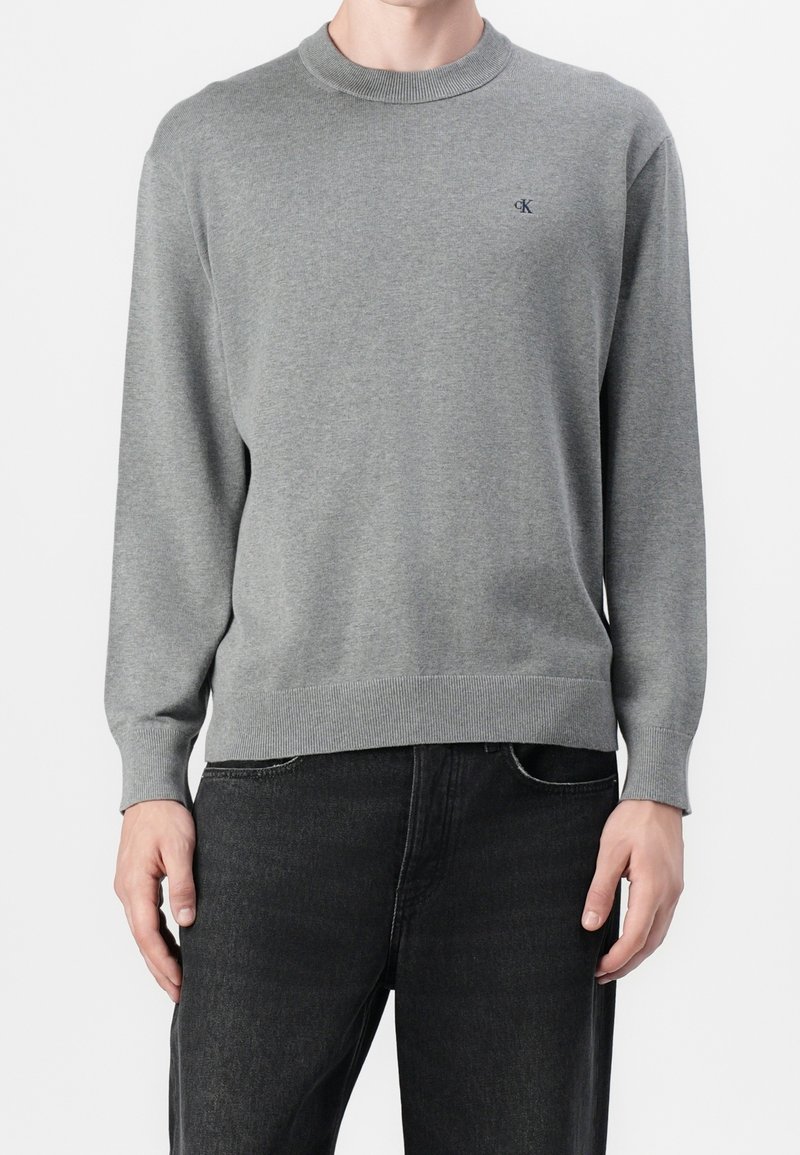 Homme portant un pull gris uni à col rond avec un petit logo "cK" sur la poitrine, associé à un jean noir, debout sur fond blanc.