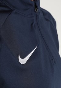 Granatowa, zapinana na suwak koszulka sportowa z białym haftowanym logo Nike na lewej piersi, z naciskiem na teksturę materiału i detalami szwów.