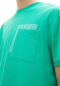 Green cotton T-shirt with short sleeves and a chest pocket. Pocket features a white text pattern. Smooth texture and standard crew neck design.