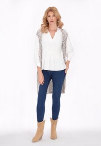 Top bianco a maniche lunghe con scollo a V, abbinato a un cardigan lungo a fantasia; indossato con jeans skinny blu e stivaletti beige.