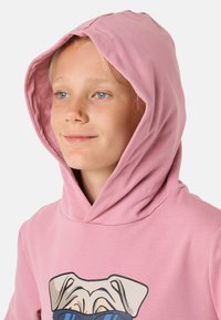 Ljusrosa hoodie med dragsko på huvan, prydd med en grafik av en hund på framsidan. Tyget ser mjukt ut med en slät textur.