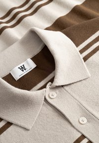 Μπεζ πόλο μπλούζα με καφέ ρίγες, πλεκτή υφή, γιακάς που διπλώνει και δύο κουμπιά στο λαιμό. Η ετικέτα εμφανίζει το λογότυπο της μάρκας.