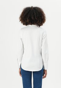 Camicia bianca a maniche lunghe con colletto posteriore, orlo leggermente curvo e polsini con bottoni, abbinata a jeans blu. Tessuto dalla texture liscia.