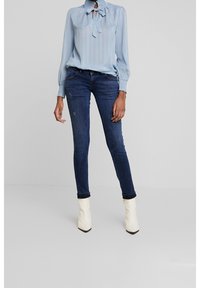 Ljusblå blus med en fluga vid halsen, långa ärmar och textilstruktur, i kombination med mörkblå skinny jeans och vita ankelkängor.