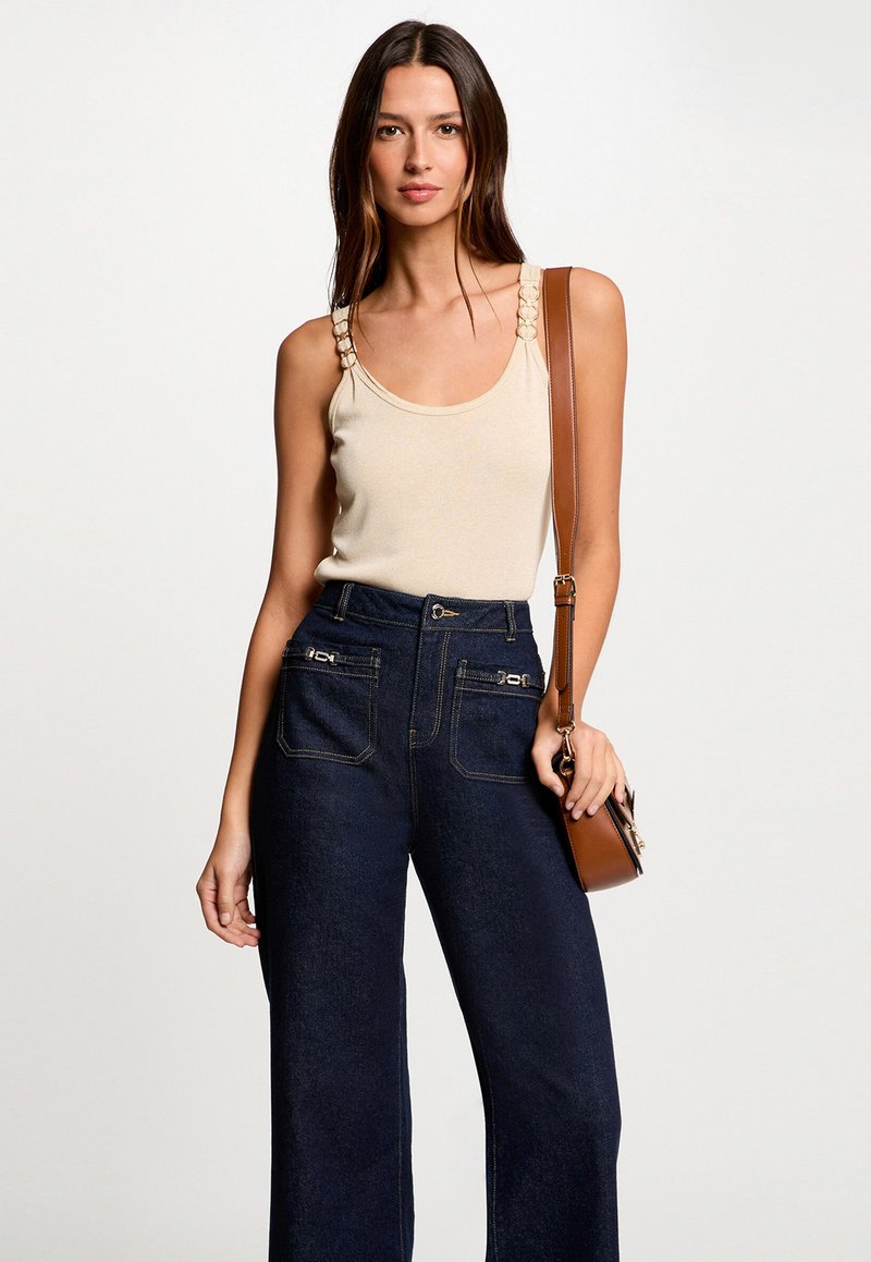 Beige sleeveless top with decorative shoulder accents paired with high-waisted dark denim wide-leg trousers. Brown crossbody bag included.