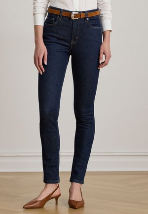 Woman wearing dark blue slim-fit jeans, a white long-sleeve shirt, brown leather belt, and brown low-heeled slingback shoes.