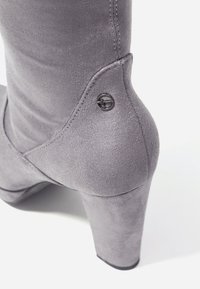 Bota de tacón alta gris hecha de un tejido suave y mate; presenta una forma curva y un pequeño acento de logo circular negro cerca del tobillo.