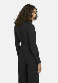Zwart jumpsuit met een getailleerde pasvorm, lange mouwen en een ceintuur in de taille. De stof lijkt glad met een minimale textuur en heeft een klassieke kraag.
