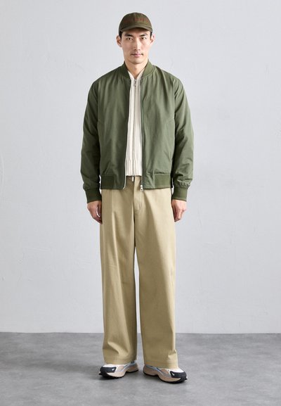Chaqueta bomber verde sobre un suéter de canalé crema, pantalones de pierna ancha beige y zapatillas negras con acentos azules. El modelo se encuentra de pie contra un fondo neutral.