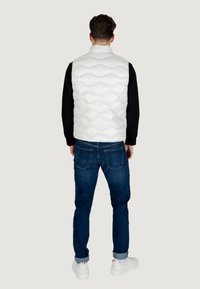Hvid polstret vest med quiltet tekstur, høj krave og lynlåslukning, parret med mørke jeans og hvide sneakers, set fra ryggen.
