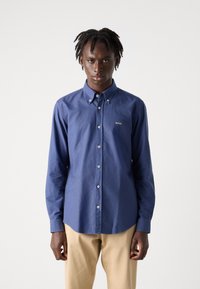 Blauw overhemd met lange mouwen en een button-down kraag, voorzien van een klein logo op de borst, gecombineerd met beige broek.