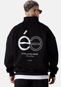 Un homme avec les cheveux courts teints et un tatouage sur le cou porte une veste noire avec un texte en français blanc et un design graphique dans le dos, tourné le dos.