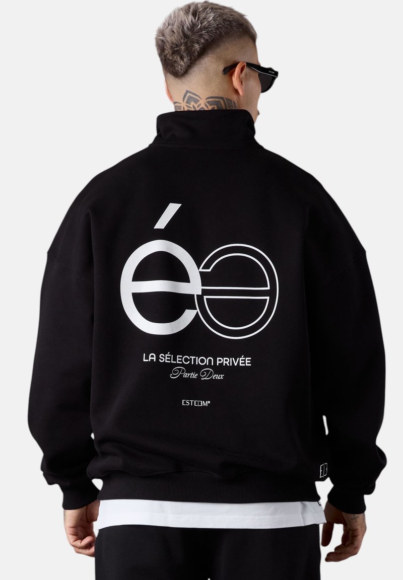 Un homme avec les cheveux courts teints et un tatouage sur le cou porte une veste noire avec un texte en français blanc et un design graphique dans le dos, tourné le dos.