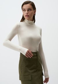 Maglione a collo alto color crema in tessuto leggero con maniche lunghe, abbinato a una gonna verde oliva con tasca laterale e chiusura frontale.