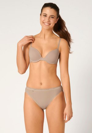 Frau mit langem, zurückgebundenem braunem Haar, die lächelt und passende beigen BH und Unterwäsche vor schlichtem hellem Hintergrund trägt.