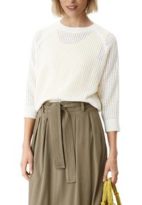 Kobieta w białym, perforowanym swetrze i wysokich spodniach taupe z materiałowym paskiem, trzymająca żółtą torbę w stylu sznurka.