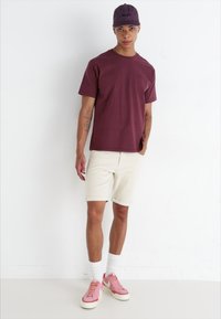 Bordeaux t-paita, beige shortsit, valkoiset perussukat ja vaaleanpunaiset lenkkarit. Lippalakki on tummanvioletti, jossa on logo. Yksinkertainen, rento design.