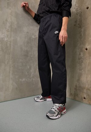 Person trägt schwarze Nike-Hose und schwarzes Shirt, steht vor einer Betonwand und trägt graue, weiße und rote Sportschuhe.