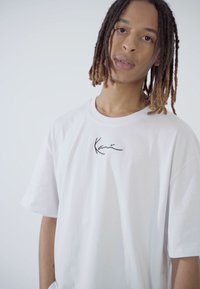 T-shirt en coton blanc avec une coupe décontractée et des manches courtes, arborant un petit logo noir en signature sur la poitrine. Texture lisse.