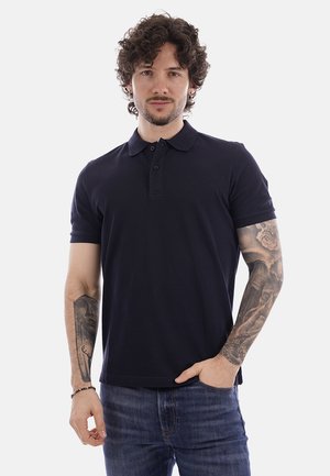 Uomo con capelli ricci che indossa una polo blu navy e jeans, mostra tatuaggi dettagliati su entrambi gli avambracci, in piedi davanti a uno sfondo bianco.