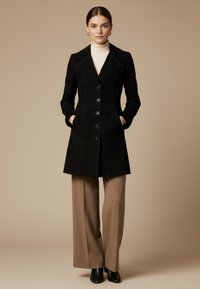 Vrouw draagt een zwarte jas met knopen over een crème coltrui, beige broek met wijde pijpen en zwarte enkellaarsjes, staand met handen in de zakken.