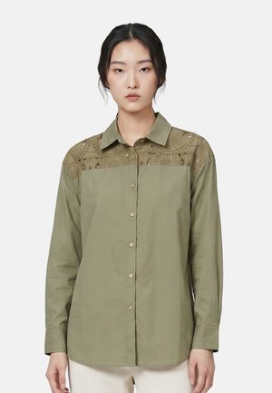 Giovane donna che indossa una camicia verde oliva a maniche lunghe con dettagli in pizzo sulla parte superiore del petto e sulle spalle, in piedi davanti a uno sfondo semplice.
