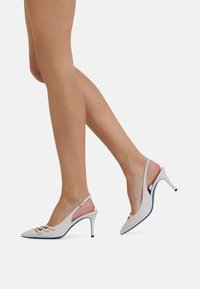 Décolleté bianchi in pelle con punta appuntita, suola blu e accessori in metallo dorato. Presenta un tacco stiletto sottile e una cinturino regolabile.