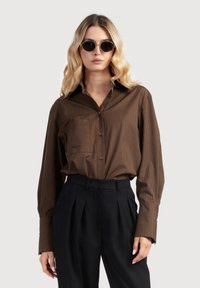 Hnedá pruhovaná košeľa s golierom na gombíky, náprsné vrecko a hnedé gombíky, kombinovaná s čiernymi nohavicami s vysokým pásom. Hladká textúra látky.