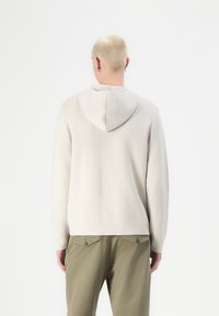 Ljusgrå hoodie i mjukt material, med rundad huva och långa ärmar. Buren med olivgröna byxor, sedd bakifrån.