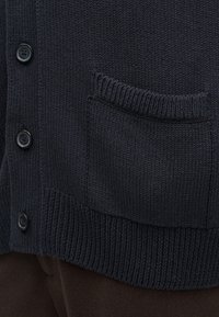 Κοντινό πλάνο μαύρου πλεκτού ζακερ με κουμπιά μπροστά και υφασμένη τσέπη πάνω από ριμπ ύφασμα στις άκρες, φορεμένο πάνω από σκούρο παντελόνι.