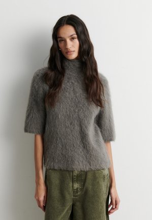 Vrouw met lang golvend haar draagt een pluizige grijze trui met korte mouwen en olijfgroene broek, staand tegen een effen witte muur.