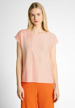 Junge Frau trägt eine hellrosa kurzärmelige Bluse und leuchtend orange strukturierte Hose, steht vor einem schlichten weißen Hintergrund.